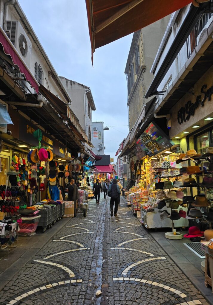 Outdoor shopping street near the Grand Bazaar lined with stalls selling hats, gloves, clothes, and souvenirs under awnings, with people walking along the cobblestone path between the tightly packed shops.
