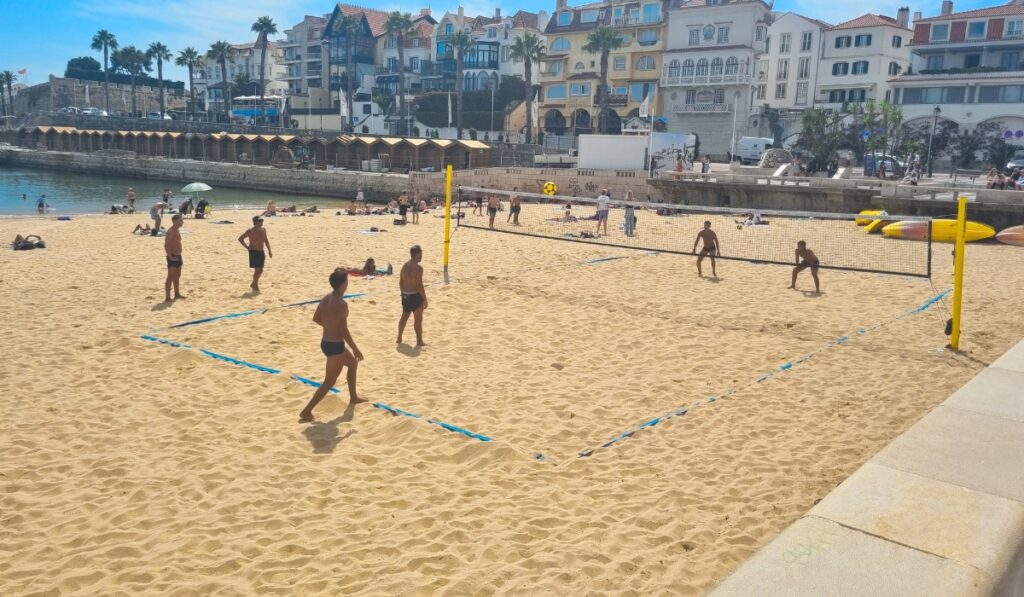 Group of men playing beach volleyball on golden sand near the water’s edge in Cascais, Portugal, with sunbathers scattered around and a backdrop of historic seaside buildings and palm trees.