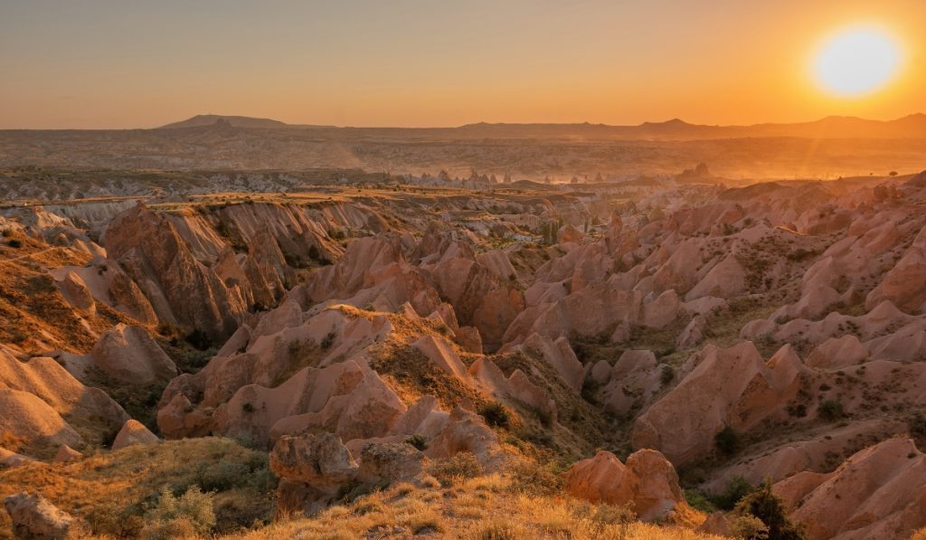 Sunset over Red Valley in Cappadocia, with jagged rose-colored rock formations glowing under the warm golden light. The landscape stretches toward the horizon as the sun sets, highlighting the valley’s dramatic ridges and soft curves.