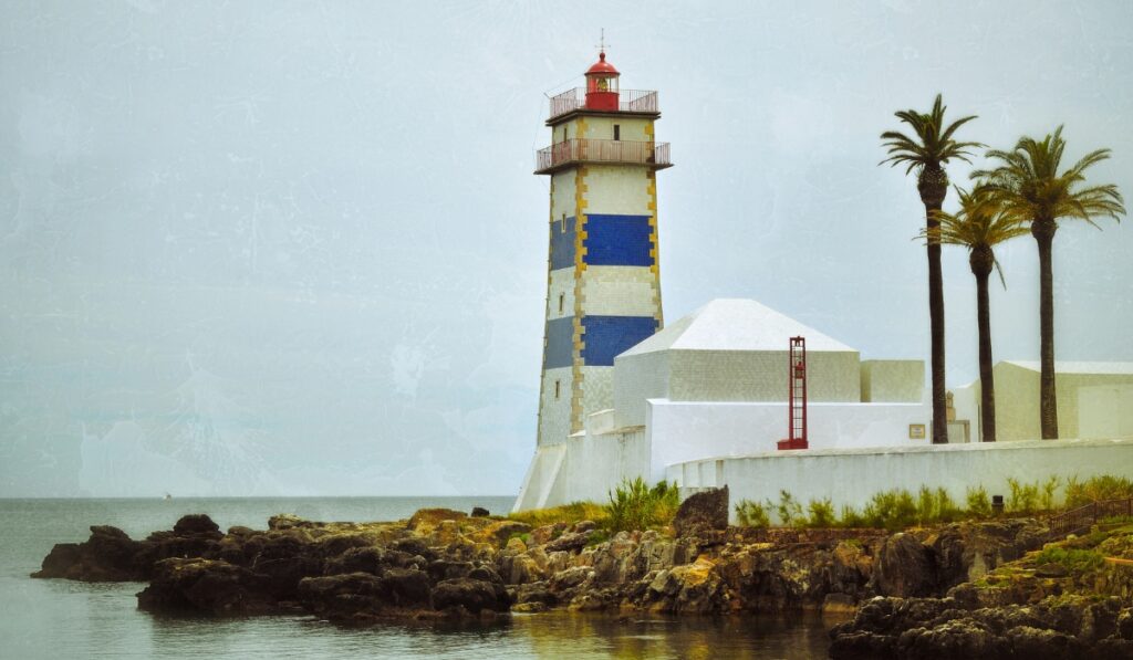 A tall coastal lighthouse with bold blue and white horizontal stripes stands near the shore, surrounded by white buildings and swaying palm trees. Rocky outcrops in the foreground meet calm water, creating a peaceful seaside scene at the Santa Marta Lighthouse.