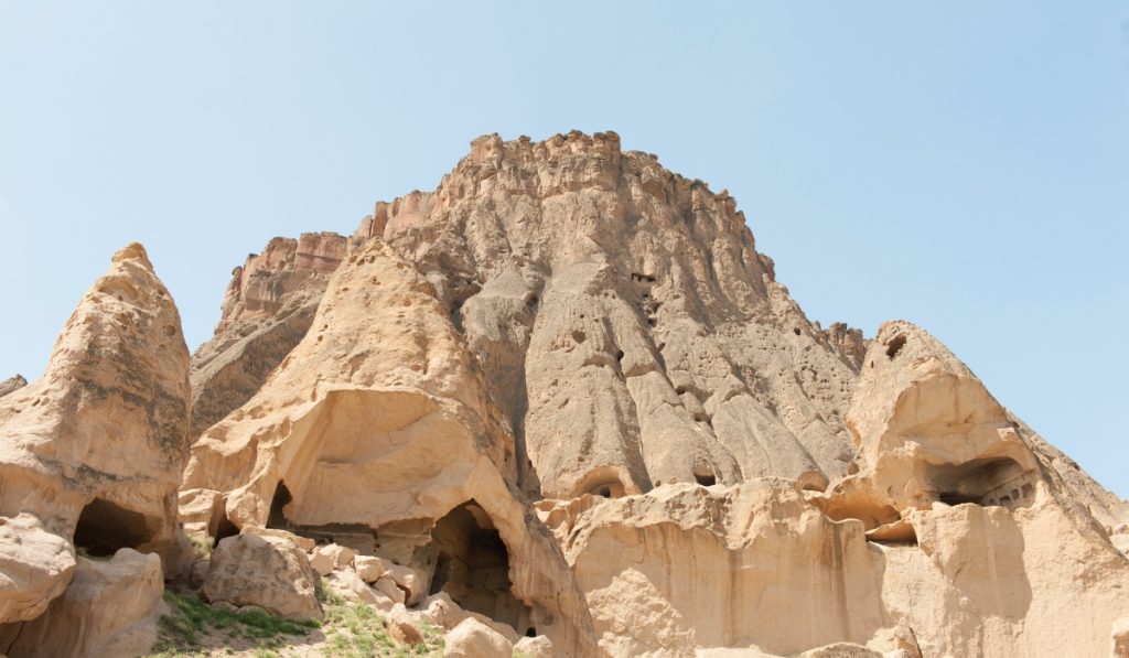 Upward view of Selime Monastery in Cappadocia, showing towering rock formations carved with ancient cave dwellings and chambers. The massive volcanic cliffs rise against a pale blue sky, highlighting the scale and rugged texture of this historic monastic site.