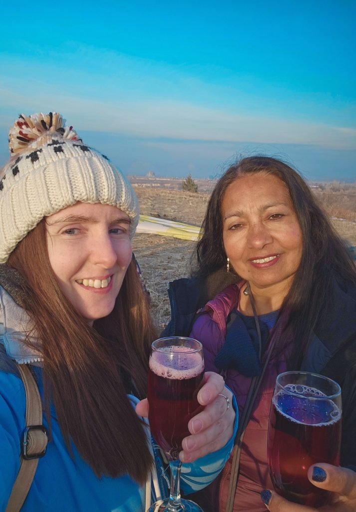 Two women smile and toast with glasses of red sparkling juice or wine, celebrating outdoors in a wide, open field under a clear blue sky. One wears a cozy knit hat and the other is dressed warmly, likely after a hot air balloon ride, with part of a deflated balloon visible in the background.