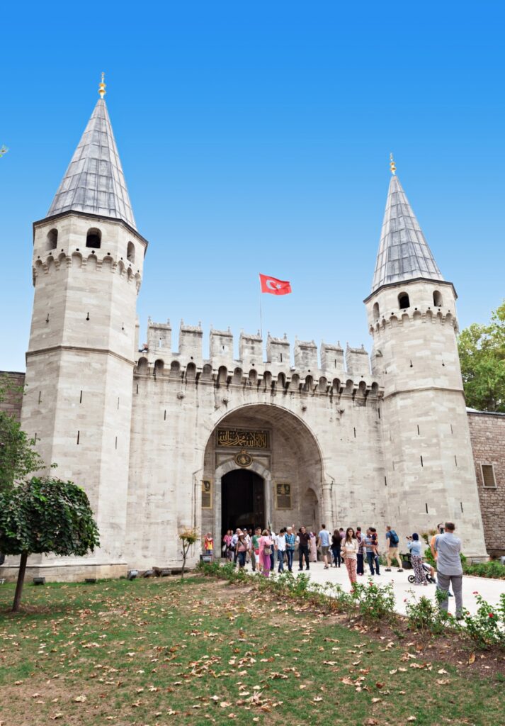 Visitors gather at the main entrance gate of Topkapı Palace, flanked by two pointed towers and topped with a Turkish flag. The grand stone gateway, known as the Gate of Salutation, stands against a clear blue sky with a path and patchy lawn in the foreground.