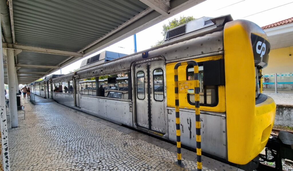 Silver and yellow CP (Comboios de Portugal) train stopped at a tiled platform under a metal roof, with a few passengers visible in the background at a Portuguese train station.