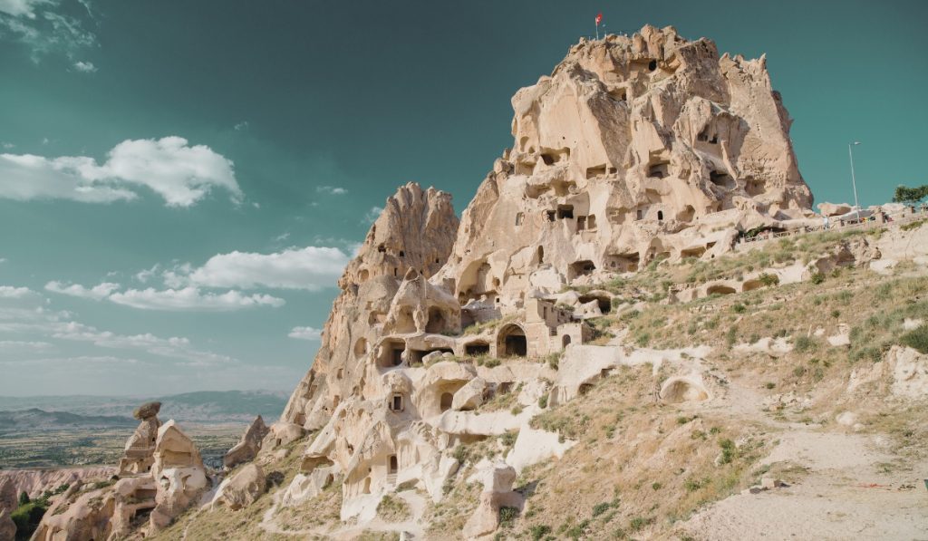 Panoramic view of Uçhisar Castle in Cappadocia, Turkey, a towering rock formation carved with dozens of cave dwellings and tunnels. A Turkish flag waves at the summit, overlooking the surrounding valleys and distant mountain ranges under a turquoise sky.