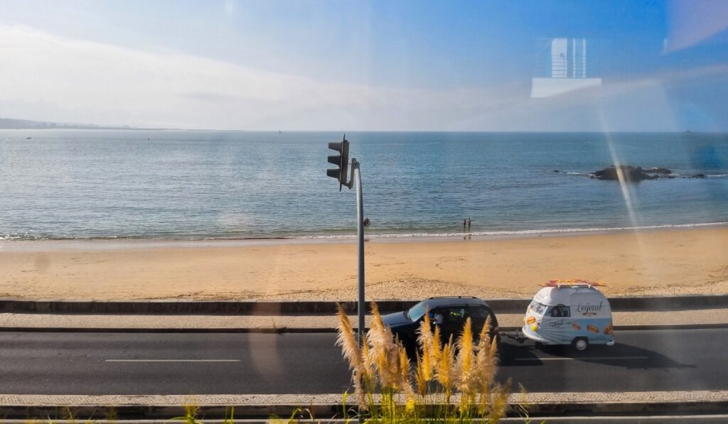 View of a calm sandy beach and the Atlantic Ocean from behind a window, with a road running parallel to the shore and a quirky food trailer decorated with hotdog graphics driving by.