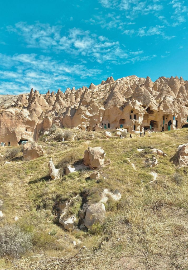 Sunlit view of Cappadocia’s Zelve Open Air Museum, showcasing a hillside of striking rock formations with carved cave dwellings and arched openings. Tourists walk among the soft tuff spires under a vivid blue sky with scattered clouds.