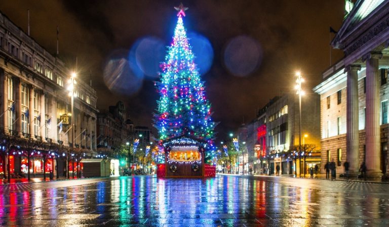A towering Christmas tree covered in multicolored lights stands in the center of a rain-slicked Dublin city street at night, reflecting vibrant colors on the wet pavement. A nativity scene is displayed at the base of the tree, surrounded by festive lights and historic buildings, creating a warm holiday atmosphere.