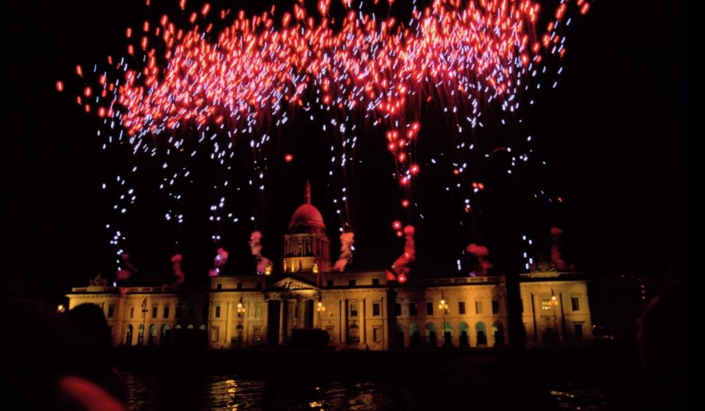 Colorful red and white fireworks burst above the illuminated dome of the Custom House in Dublin at night, reflecting on the water below.