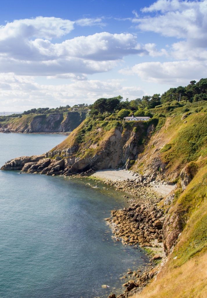 A scenic view of Dublin’s coastal cliffs near Howth, with rocky shores, calm blue waters, and a cottage nestled among lush greenery under a bright sky.