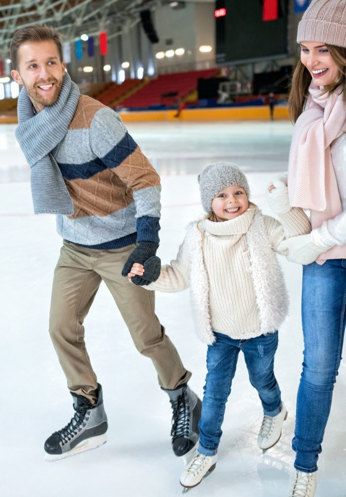 A smiling family bundled in winter clothing ice skating together indoors, with the young child joyfully holding hands with both parents.