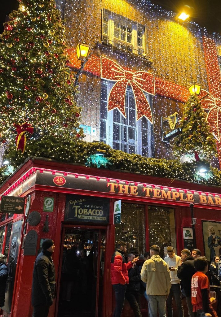 The exterior of The Temple Bar pub in Dublin is decorated with glowing Christmas lights, oversized red bows, and sparkling trees, drawing a lively crowd outside.