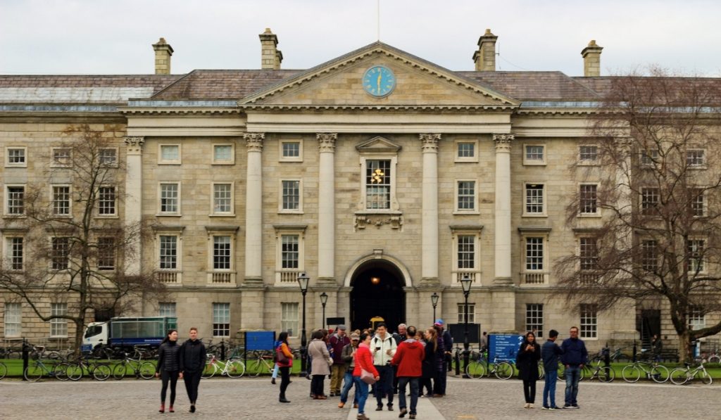 Tourists gather in front of the classical facade of Trinity College Dublin, featuring tall columns and a blue clock under a cloudy winter sky.