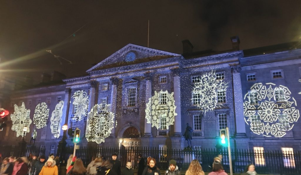 Trinity College Dublin illuminated at night with large snowflake-shaped light projections across its historic facade, drawing crowds of winter visitors.