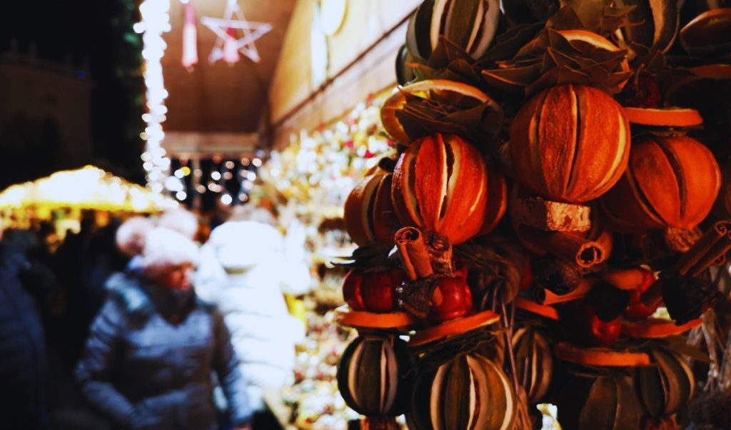 Close-up of dried orange and spice decorations with cinnamon sticks and bay leaves at a festive holiday market in Hallstatt, Austria. In the background, bundled-up shoppers browse illuminated stalls under warm string lights.
