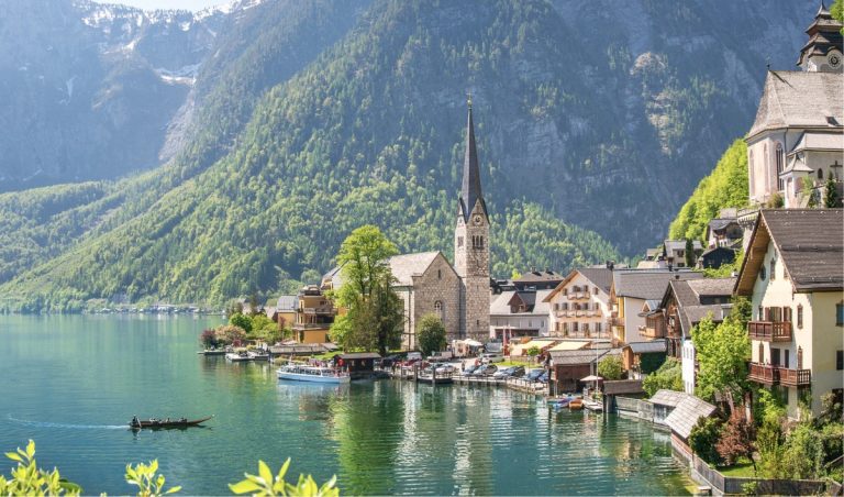 A bright springtime view of Hallstatt, Austria, ideal for a scenic day trip, with the iconic Evangelical Church and its tall spire surrounded by pastel alpine houses lining the lakeshore. A traditional wooden boat glides across the turquoise water, set against a backdrop of lush green mountains.