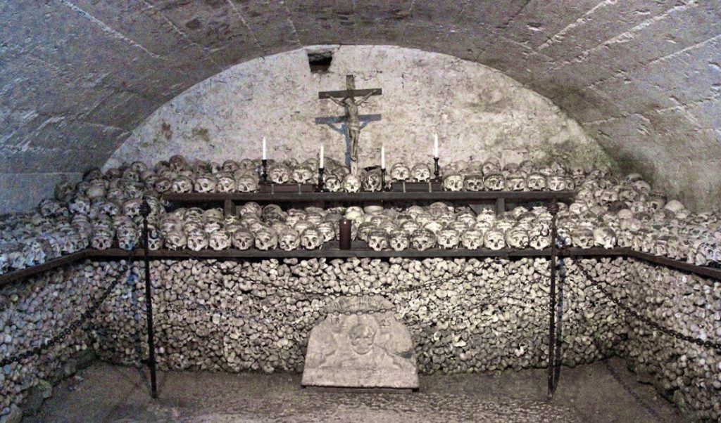 Interior of the Hallstatt Bone House (Beinhaus) featuring shelves stacked with rows of human skulls, many marked with names and dates, above a wall of neatly arranged bones. A crucifix and candles create a solemn altar at the center of the chamber.