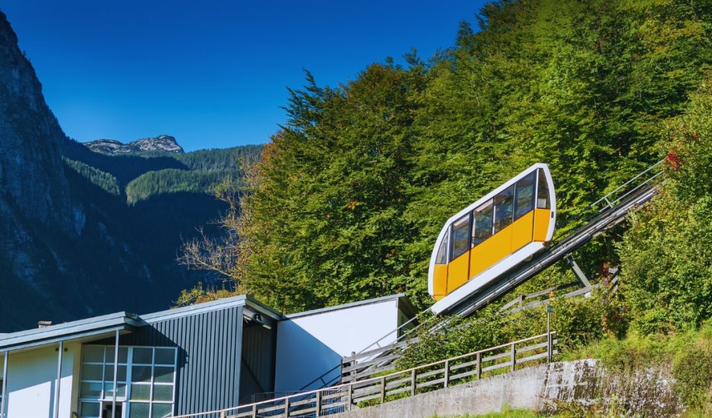 Yellow and white funicular railway car ascending a steep hillside through lush green forest near Hallstatt, Austria, with a modern station at the base and dramatic alpine peaks in the background.