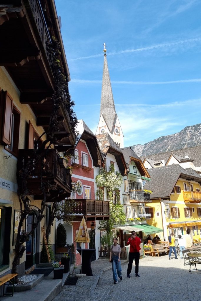 Charming street scene in Hallstatt, Austria with colorful alpine-style buildings, wooden balconies adorned with flowers, and the tall spire of the Evangelical Church rising in the background under a clear blue sky. Tourists stroll along the cobblestone path, enjoying the lively village atmosphere.
