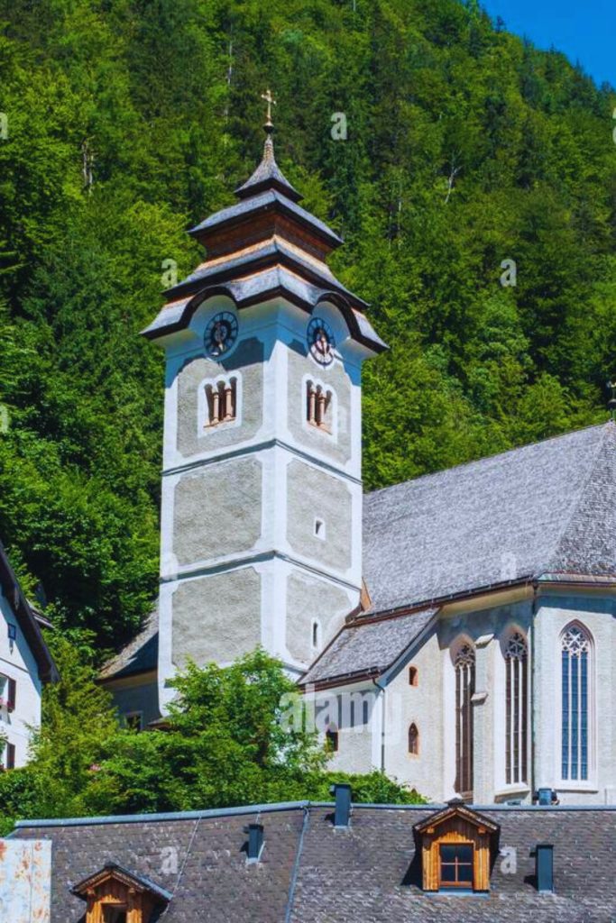 Close-up view of the Catholic Parish Church of Hallstatt, Austria, featuring its tall clock tower with arched windows and pointed roof, set against a lush green forested hillside and bright blue sky.