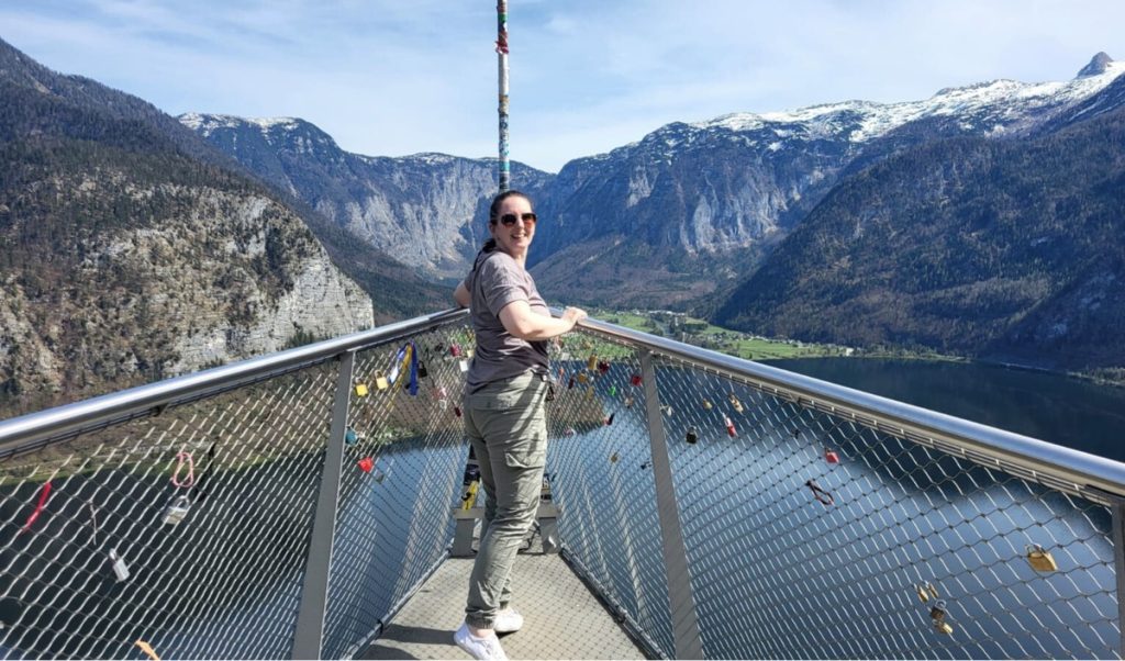 Me standing on the Hallstatt Skywalk viewing platform, smiling at the camera with a panoramic backdrop of alpine mountains, Lake Hallstatt below, and love locks attached to the metal railing.