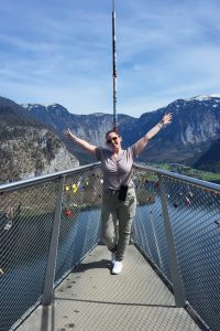 Me with arms raised stands on the Hallstatt Skywalk platform above the lake, surrounded by padlocks and panoramic alpine views.