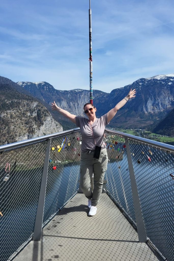 Me with arms raised stands on the Hallstatt Skywalk platform above the lake, surrounded by padlocks and panoramic alpine views.