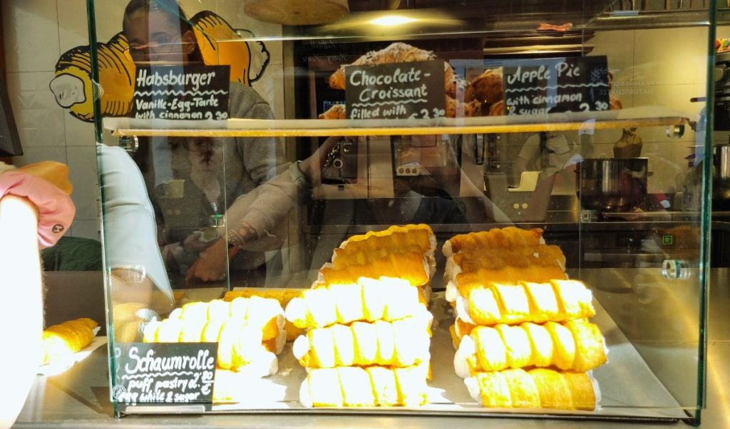 Glass pastry display case in an Austrian bakery showing stacks of golden Schaumrolle puff pastries filled with airy white cream, with other treats like chocolate croissants, apple pie, and Habsburger vanilla-egg tarts labeled on chalkboard signs above. Reflections of customers and the cozy interior are visible in the glass.