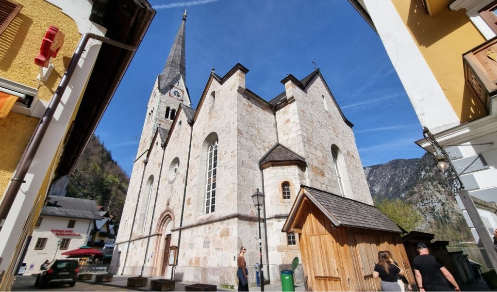Ground-level view of Hallstatt’s Evangelical Church with its Gothic-style spire, surrounded by quaint buildings and a few people enjoying a sunny day.
