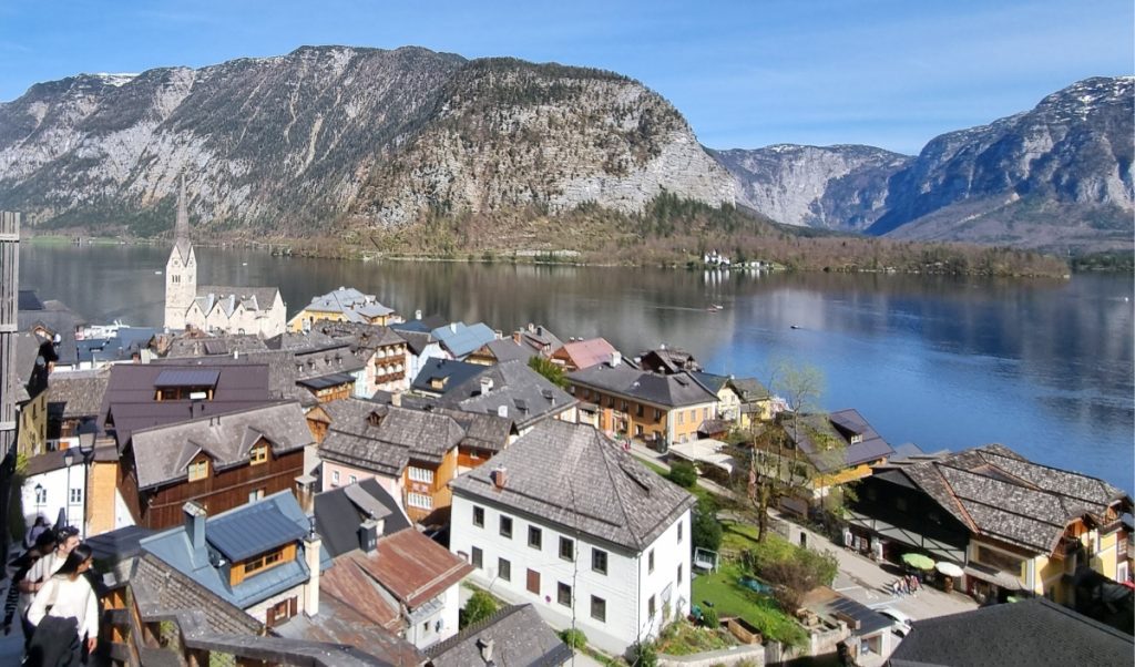 Elevated view over Hallstatt’s rooftops and lake, with pastel houses and the iconic church tower set against a dramatic mountain backdrop.
