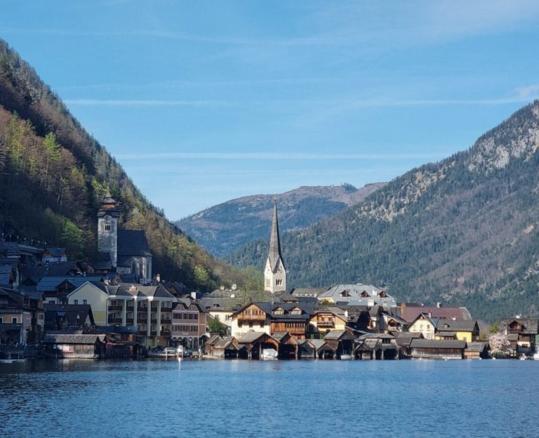 Peaceful lakeside view of Hallstatt’s waterfront buildings and twin church spires, backed by wooded slopes and blue sky.