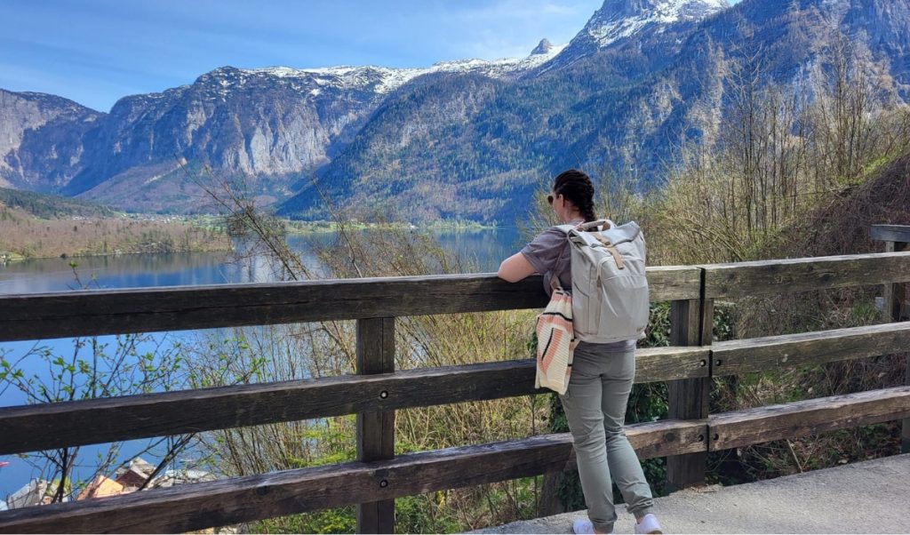 This is me with a backpack leaning on a wooden railing overlooking Hallstatt Lake and the surrounding snow-capped mountains during a sunny spring day.