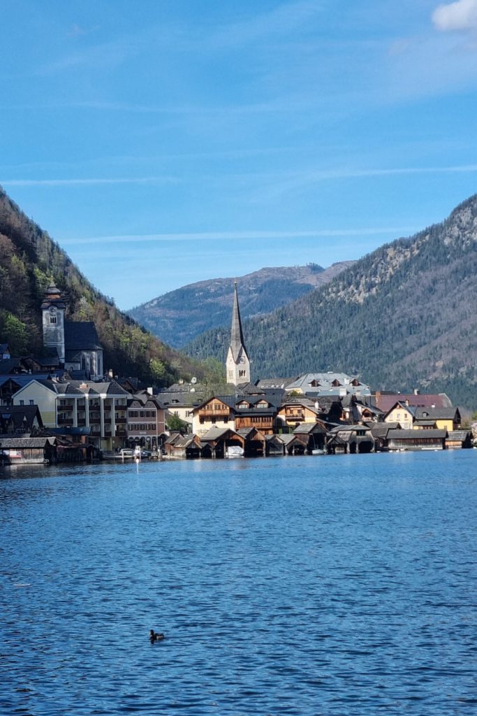Picturesque lakeside view of Hallstatt, Austria with traditional wooden boathouses lining the shore, the Evangelical Church’s pointed steeple rising in the center, and the Catholic Church’s clock tower to the left, framed by forested mountains under a clear blue sky.
