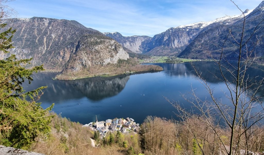 A panoramic view of Hallstatt, Austria, nestled between a serene lake and dramatic, forested mountains, with the town’s historic buildings peeking out from behind leafless trees in the foreground.