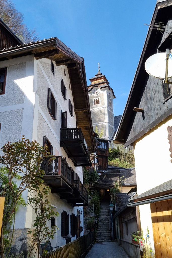Narrow alleyway in Hallstatt, Austria with traditional alpine houses featuring wooden balconies and shuttered windows, leading up a stone staircase toward the Catholic Parish Church’s clock tower under a clear blue sky.