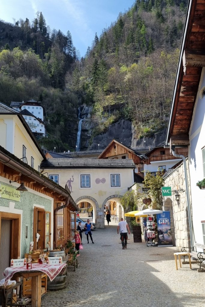 Cobblestone street in Hallstatt, Austria lined with quaint shops and signs for local schnapps and souvenirs, leading to an archway framed by alpine buildings. In the background, a narrow waterfall cascades down a forested hillside under a clear blue sky.