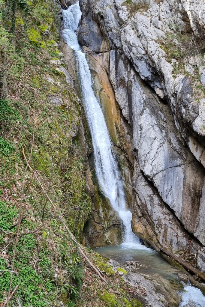 A narrow waterfall cascades down a rocky cliff surrounded by moss-covered stone and sparse spring greenery, creating a peaceful nature scene near Hallstatt.