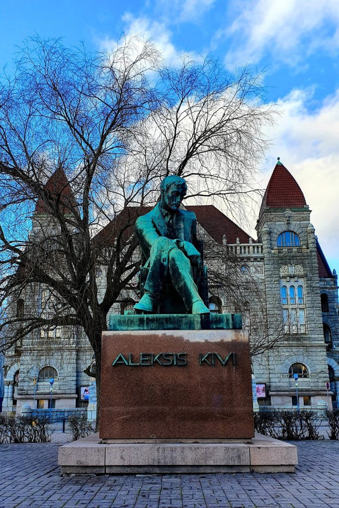 Bronze statue of Aleksis Kivi seated on a stone pedestal engraved with the text ALEKSIS KIVI, set in front of an ornate historic building and leafless trees. The monument honors the Finnish author in a central city square.