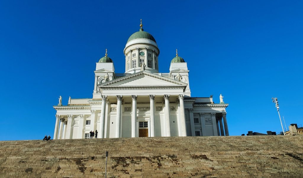 White neoclassical cathedral with tall columns and green domes stands at the top of wide stone steps under a clear blue sky. The grand façade and elevated position emphasize the scale and importance of the historic cathedral.