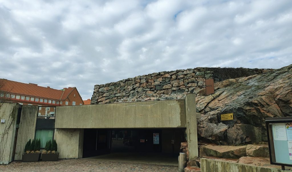 Concrete entrance built into a rocky hillside leads into the underground church, with stacked stone walls above and a cloudy sky overhead. The low modern façade blends into the natural rock landscape, emphasizing the church’s distinctive subterranean design.