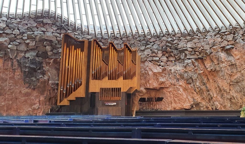 Large pipe organ with tall wooden pipes stands against a natural rock wall beneath a circular skylight ceiling. Rows of dark blue pews face the organ, highlighting the unique stone interior of the modern church carved into rock.