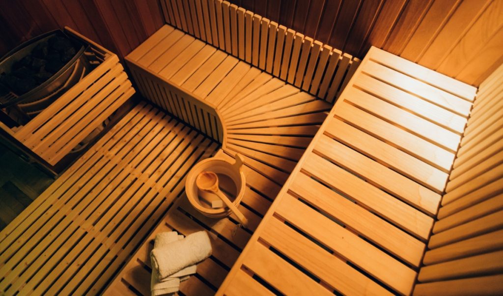Warm wooden sauna interior with tiered bench seating and soft amber lighting creating a relaxing spa atmosphere. A wooden bucket with ladle and neatly folded white towels rest on the bench, highlighting the traditional sauna setup and inviting self care experience.