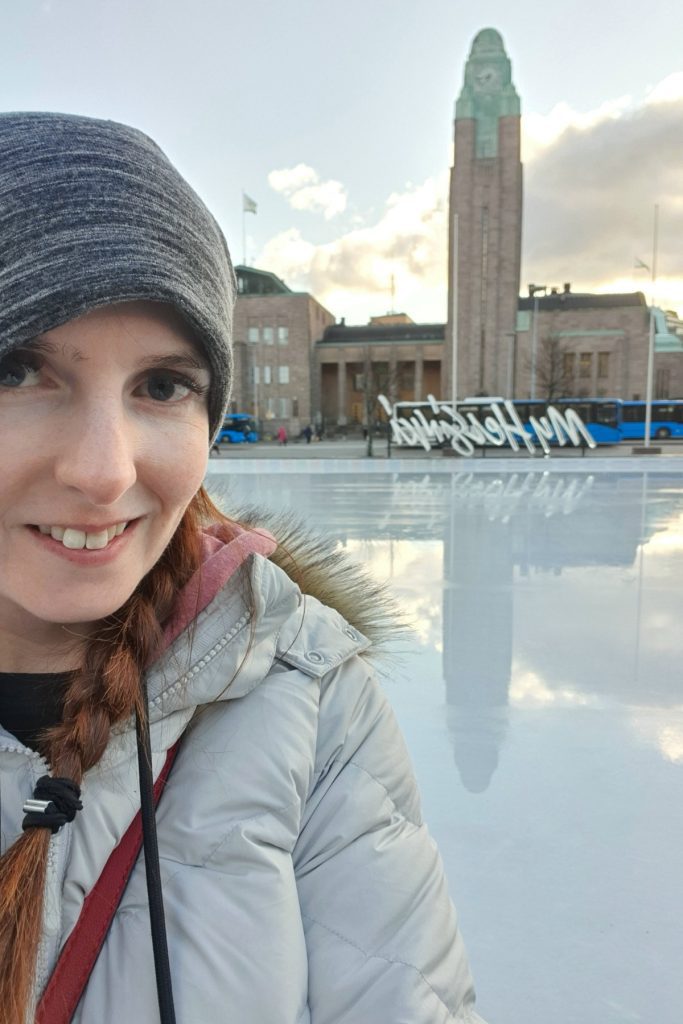 Me in a gray knit hat and light winter coat takes a selfie in front of a large building with a tall clock tower at sunset. The icy plaza reflects the architecture and the white My Helsinki sign in the background.