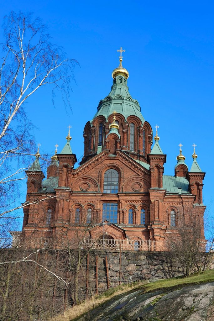 Red brick cathedral with green copper domes and gold crosses rises against a vivid blue sky, framed by bare tree branches in the foreground. The ornate architecture and hillside setting highlight the historic church as a striking city landmark.
