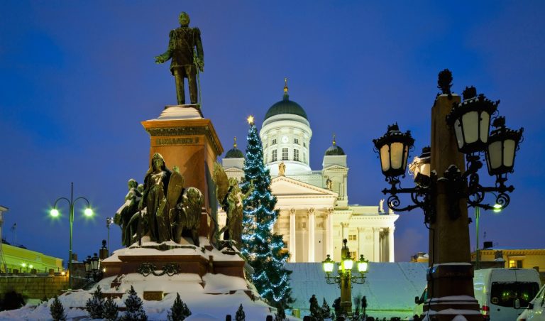 A snow covered statue stands in the foreground of Helsinki Cathedral at dusk, with a lit Christmas tree glowing in front of the white neoclassical church and its green domes. Ornate street lamps frame the scene as soft blue evening light highlights the winter atmosphere in Senate Square.
