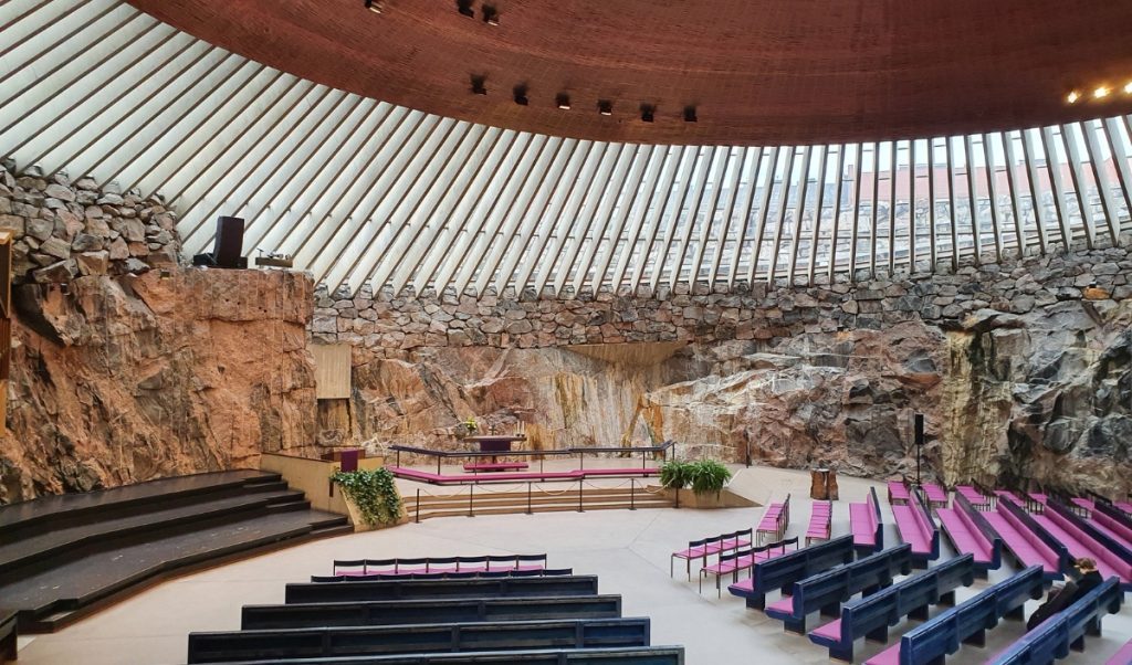 Wide view of a rock carved church interior with exposed stone walls and a round skylight dome letting in natural light. Simple altar with greenery and pink cushioned seating create a calm, contemporary worship space surrounded by raw granite.
