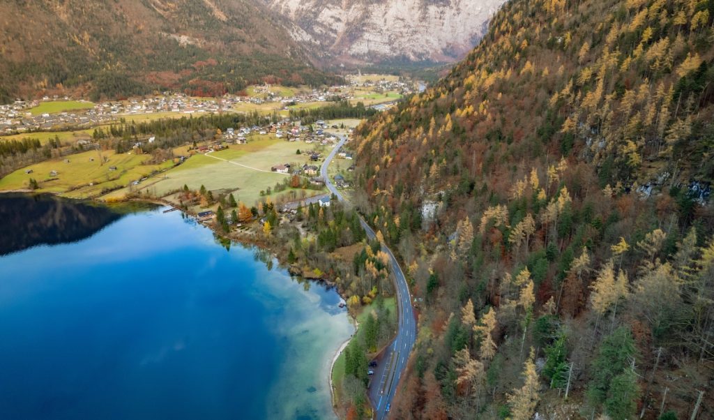 Aerial view of Obertraun, Austria with a winding road running between a deep blue alpine lake and forested mountainside in autumn colors. Scattered homes and green fields stretch across the valley beneath rugged, rocky peaks.