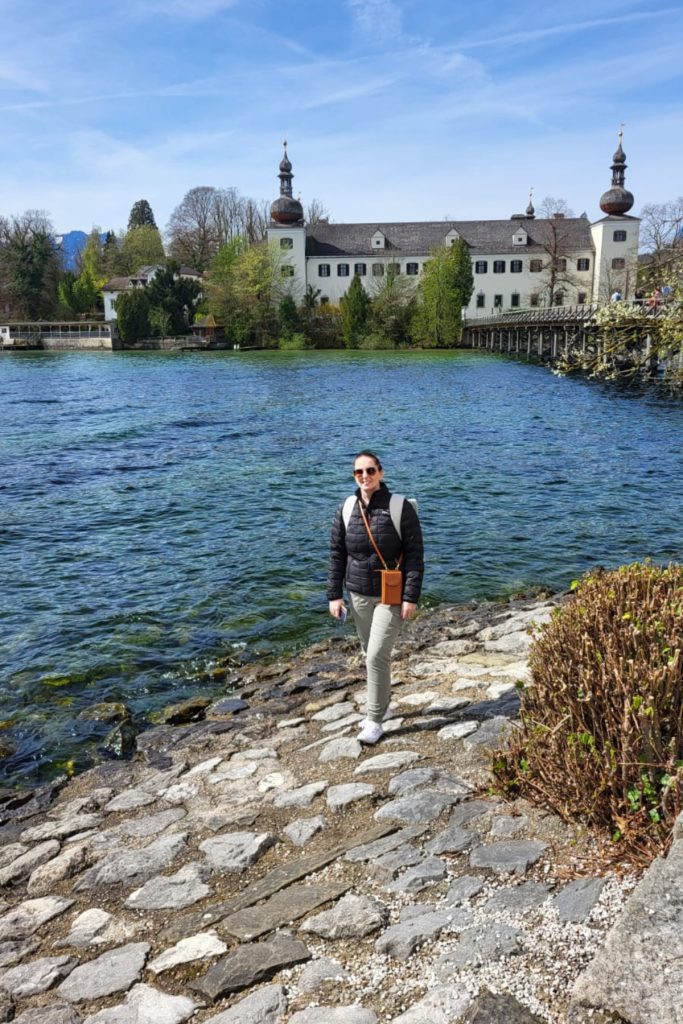 Me smiling by a rocky lakeshore with crystal-clear blue water in the foreground and Seeschloss Ort, a historic Austrian lakeside castle with onion-domed towers, in the background. A long wooden bridge leads to the castle, framed by spring greenery under a bright blue sky.
