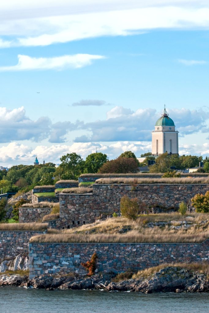 Historic stone fortress walls covered in grass rise above the waterfront under a bright blue sky with scattered clouds. A domed church with a green copper roof stands in the background, adding architectural contrast and highlighting the coastal city landscape.