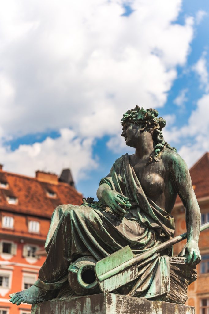Bronze statue of a seated woman with a weathered green patina, holding a paddle and looking to the side against a bright sky. Red roofed historic buildings blur in the background, drawing focus to the sculpture’s detailed drapery and calm pose.